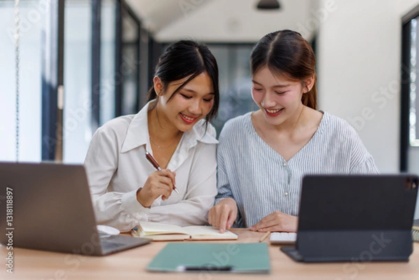 Fototapeta Happy professional young women collaborating with enthusiasm in a modern office setting. Two asian female entrepreneurs using a laptop for a productive business meeting in a corporate workplace.
