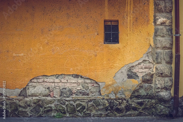 Fototapeta An old residential building wall in Szentendre, Hungary, with peeling plaster revealing brick and stone layers. A small window adds character. The textured facade shows age, history, and urban decay