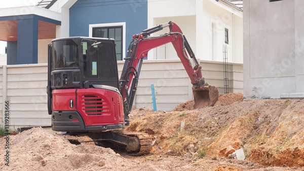 Obraz A small red excavator is digging a hole to prepare for building a wall around a concrete house on background Concept of working with machinery at a construction site.