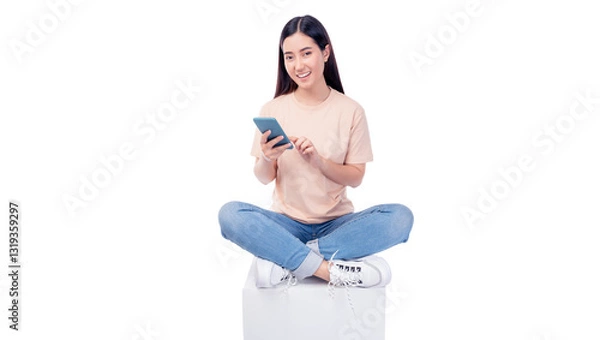 Fototapeta Cheerful young Asian woman sitting cross-legged on a white cube, engaging with smartphone. She wears T-shirt and blue jeans, smiling while interacting with the phone isolated on white background