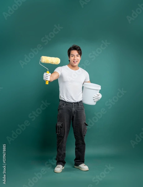 Obraz A painter in a white shirt, black pants, and white gloves holds a paint roller and bucket against a solid green background, ready to paint a room.