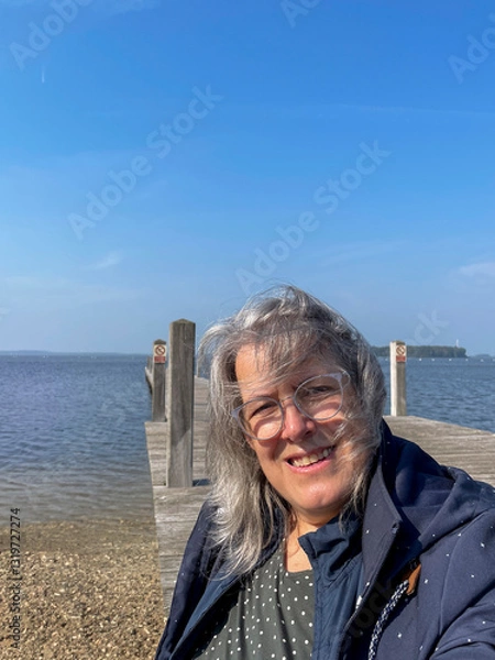 Obraz Frau mit langen grauen Haaren sitzt am Steg am Meer, ihre Haare sind vom Wind zerzaust