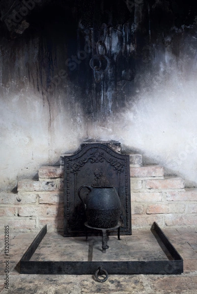 Obraz Rustic old fireplace with a blackened iron kettle on a stand. Soot-covered walls and antique brickwork create a historical, vintage atmosphere. Warm tones contrast with dark, aged metal elements.