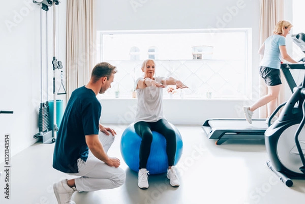 Fototapeta Elderly woman performing balance exercises on a stability ball under supervision of a male trainer in a bright, modern gym setting