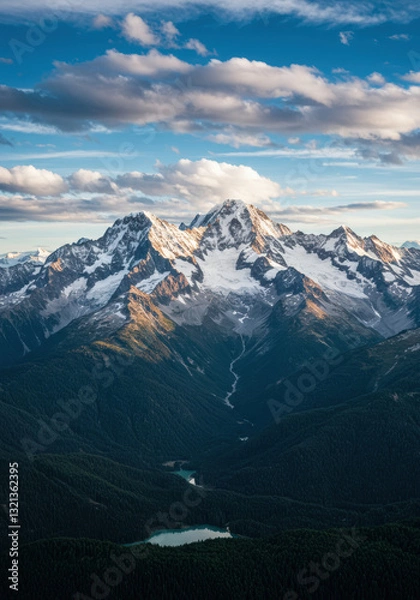 Fototapeta Majestic snow-capped mountains rise above a dense forest under a sky of floating clouds.
