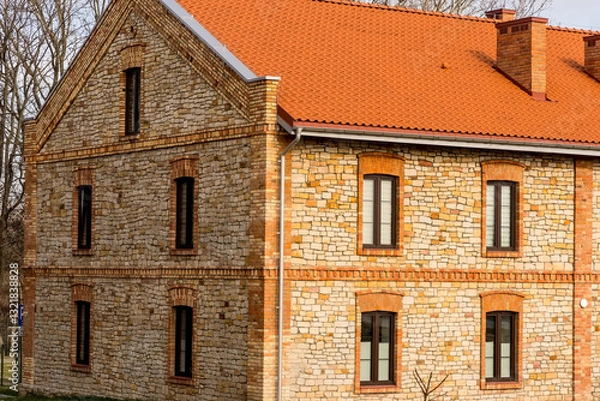 Fototapeta Piękny zabytkowy kamienny dom świeżo odnowiony, w pochmurny lutowy dzień z ciepłą beżową fasadą, zaakcentowaną czerwoną cegłą wokół okien i krawędzi. Zwieńczony żywym czerwonym dachem z dachówki.