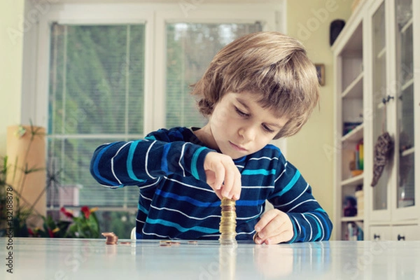Fototapeta Little boy making stack of coins, counting money at table. Learning financial responsibility and planning savings concept.
