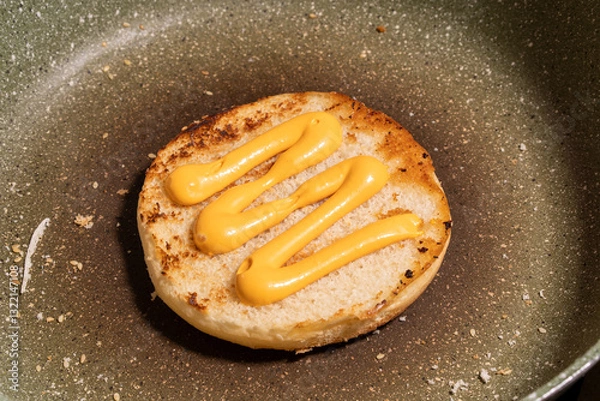 Fototapeta A sesame seed bun with zigzag mustard lines rests on a skillet, surrounded by crumbs and sesame seeds, ready for burger assembly.