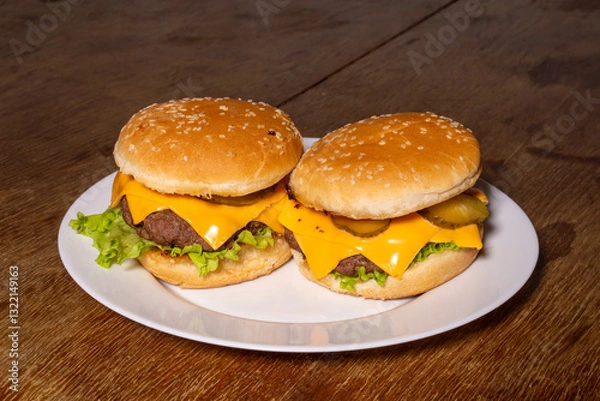 Fototapeta Two cheeseburgers with beef patties, melted cheese, lettuce, and pickles on sesame buns, served on a white plate atop a rustic wooden surface.