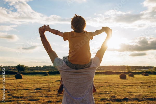 Fototapeta Father carrying son on shoulders in a sunny countryside field, holding hands and enjoying a joyful summer moment