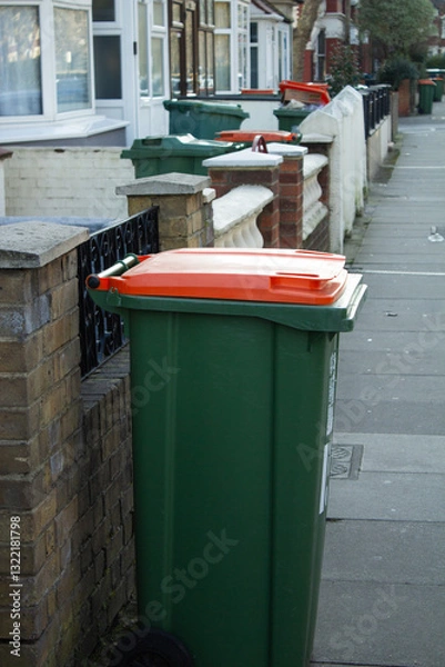 Fototapeta A green trash bin with an orange lid for recyclable waste stands on the street in the city, with buildings and other trash bins in the background.