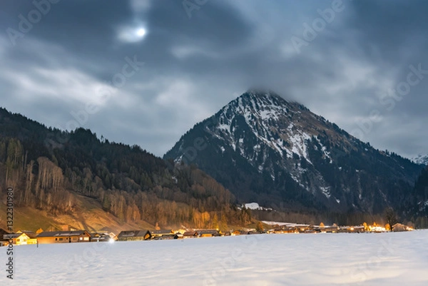 Fototapeta Winter night landscape in the mountains, full moon hidden behind clouds over the mountains. Small village in the Alps, houses lit at night, view from a hiking trail.at night, view from a hiking trail.