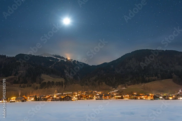 Fototapeta Winter night landscape in the mountains, full moon in the starry sky above the mountains. Small village in the Alps, houses illuminated at night, view from a hiking trail.