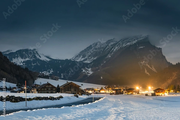 Fototapeta Winter night landscape in the mountains, snow-covered foothills with a small village in the Alps, houses illuminated at night, view from a hiking trail.