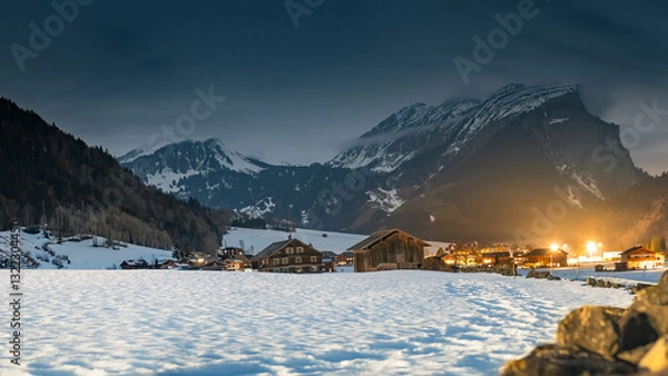 Fototapeta Winter night landscape in the mountains, snow-covered foothills with a small village in the Alps, houses illuminated at night, view from a hiking trail.