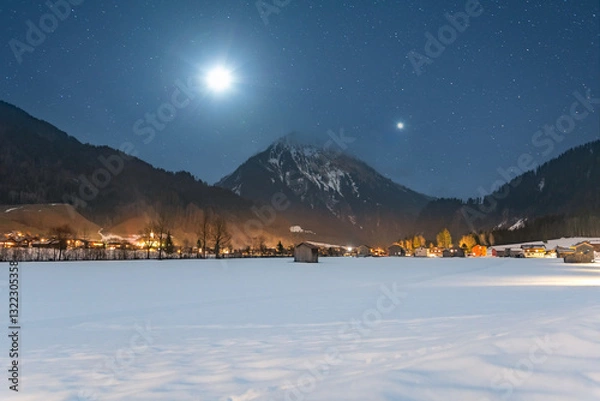 Fototapeta Winter night landscape in the mountains, full moon and planet Venus in the starry sky above the mountains. Small village in the Alps, houses illuminated at night, view from a hiking trail.