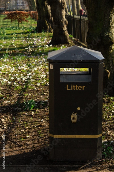 Fototapeta A black trash bin located in the park, with trees and flowers in the background.