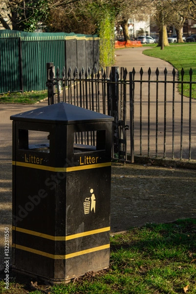 Fototapeta A black trash bin located in the park, with gates and a pathway in the background.