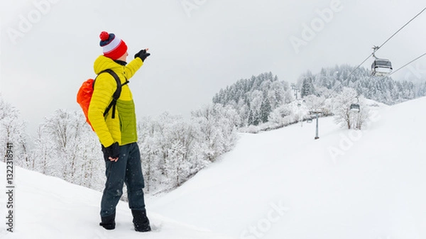 Fototapeta A young man stands on the trail and shows how high the gondola lift goes. Winter landscape on a mountain hiking trail on a cloudy and foggy day.