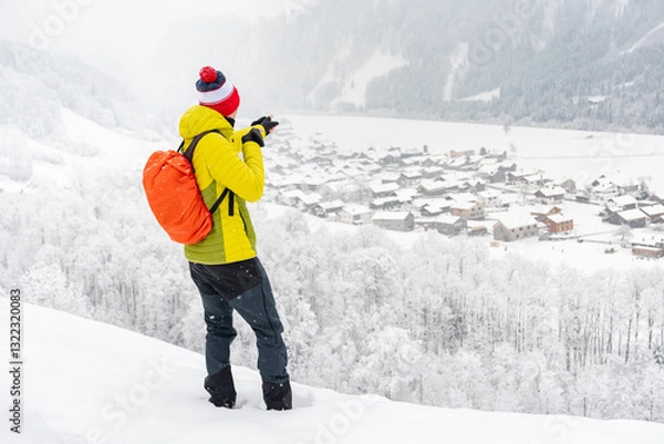Fototapeta A young man stands on a trail and looks at a town in a mountain valley. Winter landscape on a mountain hiking trail on a cloudy and foggy day.