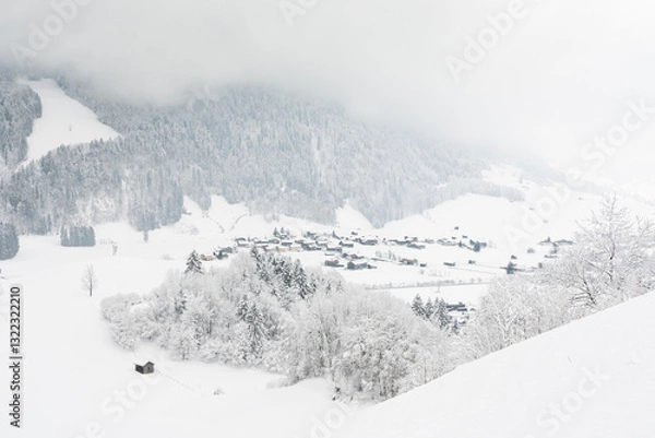 Fototapeta View of a small town in the Alps in a snowy mountain valley. Winter landscape with a view of a mountain hiking trail on a cloudy and foggy day.