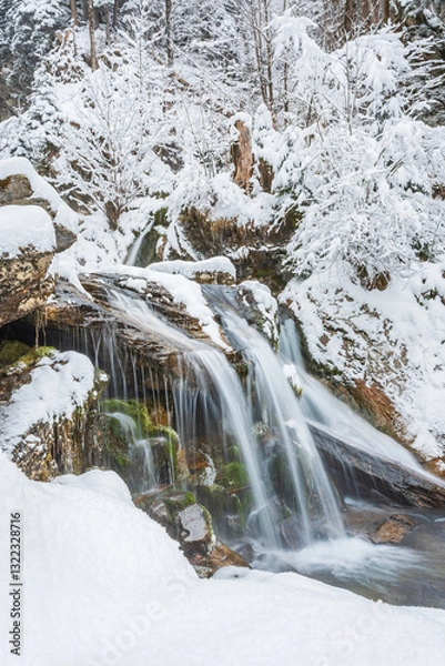 Fototapeta Small waterfall on a mountain river, water flows down snow-covered rocks. Winter landscape on a mountain hiking trail.