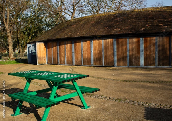 Fototapeta Green benches in the park with attached boards for playing chess, with a wooden building and trees in the background.