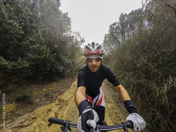 Obraz POV, Original point of view. Happy Young man on bicycle take a selfie with action cam and selfie stick. Training on mountain bike on a mountainous road in a rainy day
