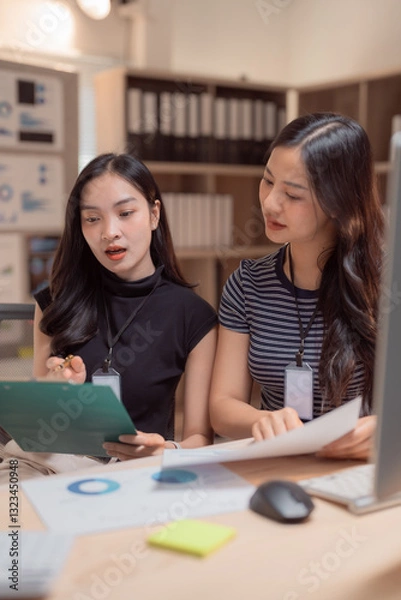 Fototapeta Two young businesswomen are discussing financial data in a modern office, collaborating on documents and analyzing charts to make informed business decisions