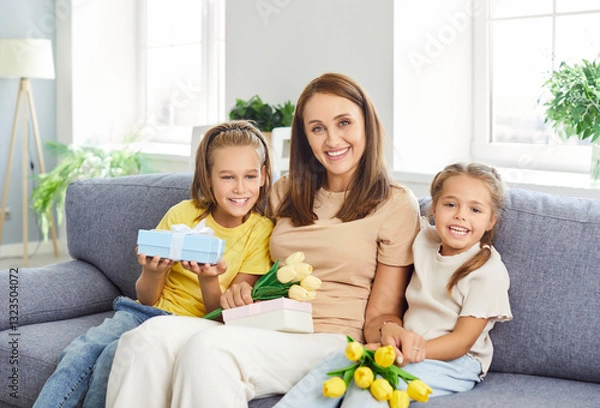 Fototapeta Portrait of a smiling children congratulating happy young mother with flowers bouquet of tulips and a present box sitting on the sofa at home. International Women's or Mothers Day concept.