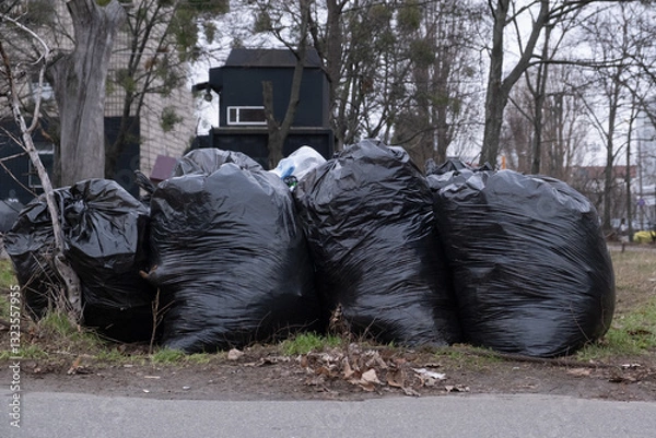 Fototapeta In a neglected urban park area, a cluster of black garbage bags accumulates beside trees, highlighting the ongoing challenges related to waste management in our communities and urban environments