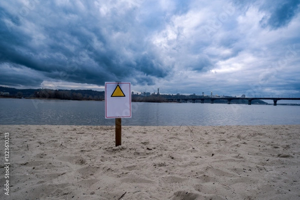 Fototapeta A cautionary sign stands prominently in the sandy foreground as stormy clouds ominously loom over a tranquil river: Embankment of the Dnipro River in Kyiv