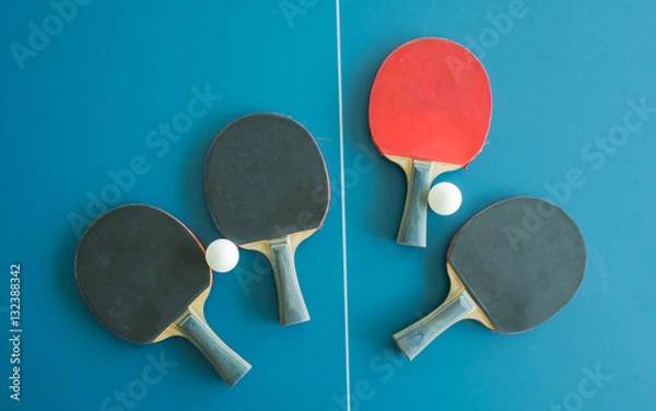 Obraz A red table tennis bat surrounded by three black table tennis bats and balls on a table with white vertical line. Concept of uniqueness, outstanding.