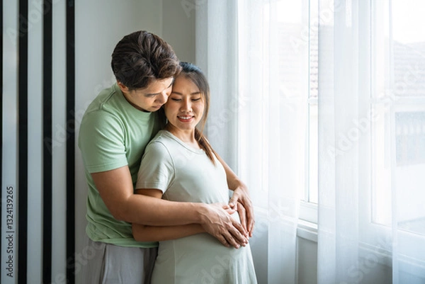 Obraz Romantic Asian husband embracing pregnant wife near window with soft natural light
