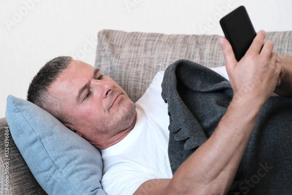 Fototapeta A Relaxed Man Comfortably Using His Smartphone While Seated on the Cozy Couch with a Blanket