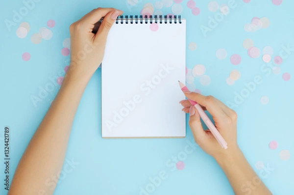 Fototapeta Well-groomed female hands with a pen near a notebook on a blue background. To-do list. Notes. Blank sheet for text.