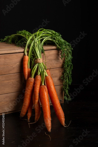 Fototapeta Close-up of a bunch of carrots with green leaves on a wooden crate, black background, vertical with copy space