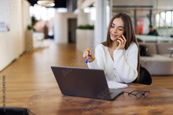 Fototapeta Cheerful young beautiful woman talking on mobile phone and using laptop with smile while sitting at her working place
