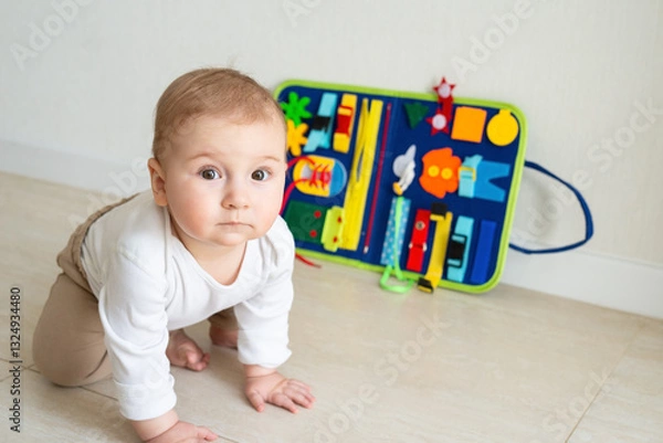 Fototapeta Baby girl plays on the floor. On the background developing bright handmade toy for fine motor skills made of felt. Early childhood development, soft busy book with with laces, zippers