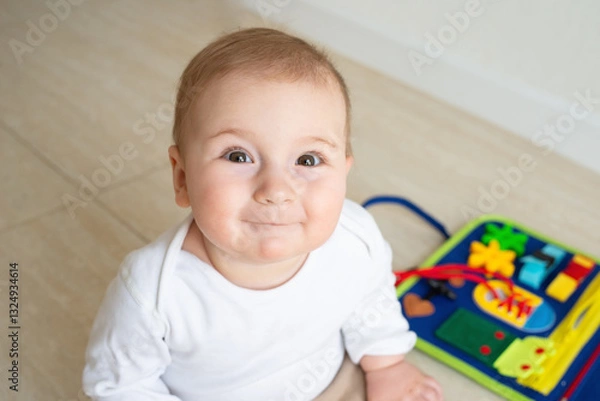 Fototapeta Cute baby girl smile sitting on the floor. On the background developing bright handmade toy for fine motor skills made of felt. Childhood development, soft busy book with laces, zippers and Velcro.