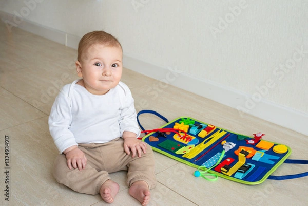 Fototapeta Cute baby girl smile on the floor. On the background developing bright handmade toy for fine motor skills made of felt. Early childhood development, soft busy book with with laces, zippers and Velcro.
