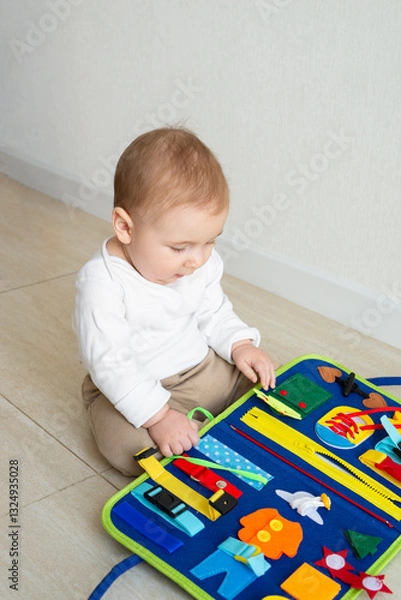 Fototapeta Baby girl sit plays on the floor with a developing bright handmade toy for fine motor skills made of felt. Early childhood development, soft busy book with with laces, zippers and Velcro