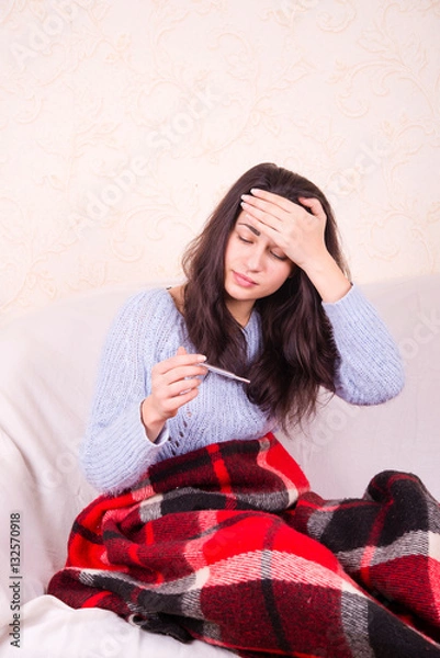 Fototapeta girl on the couch under a blanket holding a thermometer