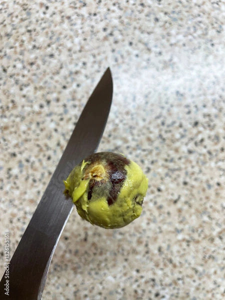 Fototapeta Round brown avocado pit with green avocado pieces attached to gray metal sharp blade of large knife on tabletop background, close-up, top view. Removed avocado pit. Cooking tricks.