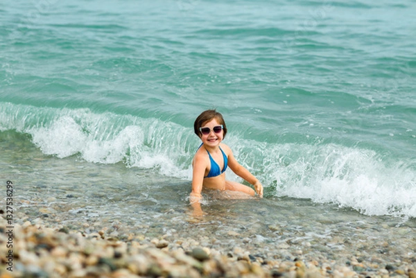 Obraz A small, beautiful girl in a blue swimsuit and sunglasses, swimming on the beach