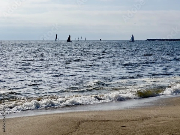 Fototapeta Silhouetted sailing boats in the distance, on Pacific Ocean in Southern California, with waves breaking on sandy beach in foreground. Light clouds overhead