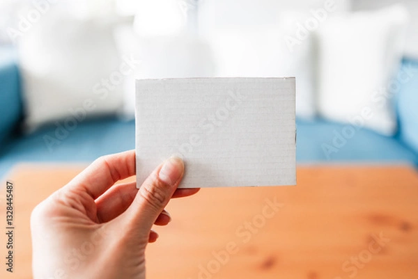 Fototapeta hand holding small cardboard label with blank copy space in front of stylish sofa and coffee table