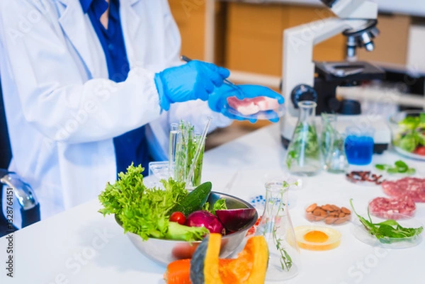 Obraz Middle-aged female laboratory worker wearing blue gloves conducts lab test on food ingredients lettuce, cucumber, pork. Using microscope, she analyzes nutrients, preservatives, food safety records.