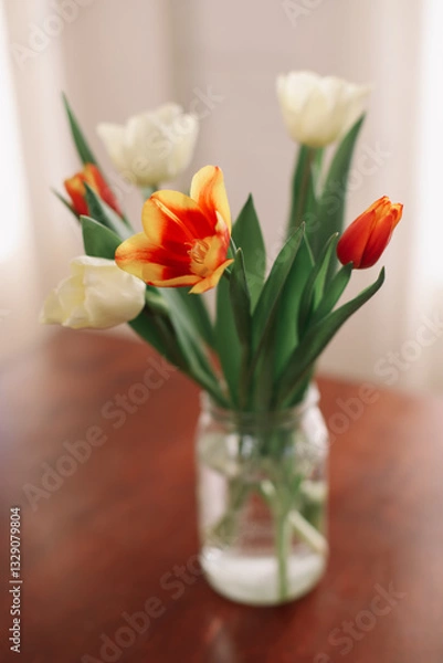 Fototapeta Bouquet of beautiful fresh tulips in a glass vase on wooden table in cozy modern interior. Spring flowers in glass vase at home.