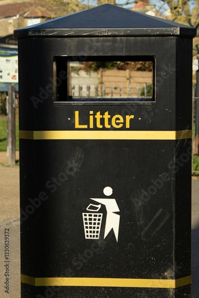 Fototapeta A black trash bin with yellow stripes in the park.